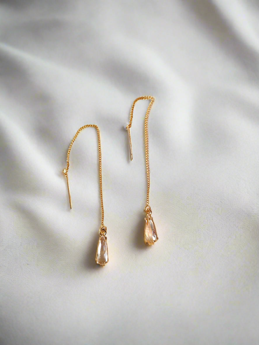 A pair of gold-plated threader earrings with a crystal pendant, displayed against a white fabric background.